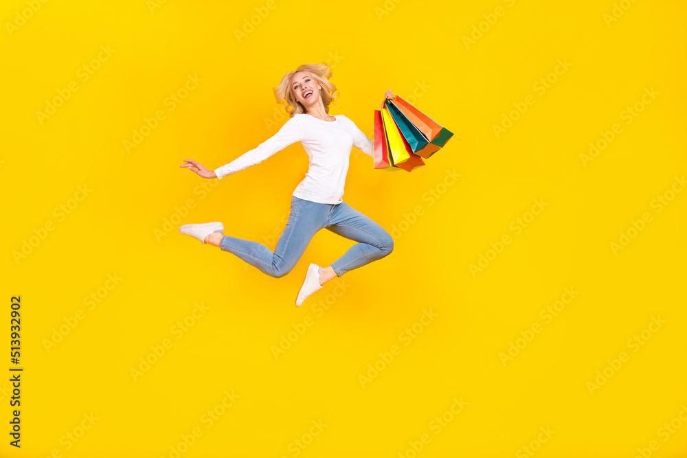 Full body photo of nice young blond lady jump with bags wear shirt jeans footwear isolated on yellow background