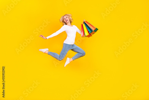Full body photo of nice young blond lady jump with bags wear shirt jeans footwear isolated on yellow background © deagreez