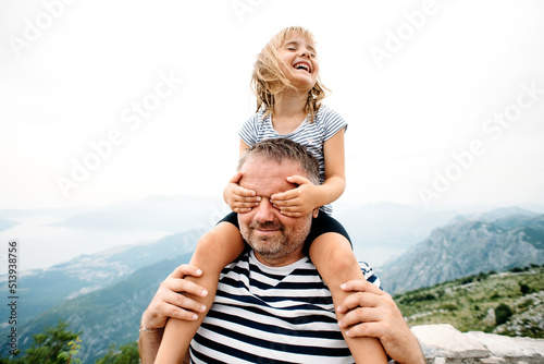 dad and daughter smiling and having fun together. daughter sitting on dad's shoulders and covers dad's eyes with hands. They are standing on the view point on Kotor bay, Montenegro. Wide angle photo