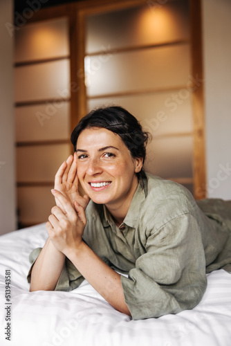 Cheerful young caucasian girl smiling looking at camera lying on bed in morning. Brunette wears casual clothes relaxing on weekends. Relaxation concept