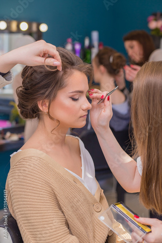 Women's hands paint the girl's eyes and curl her hair in the salon. Make-up artist and hairdresser create an image of a young woman. Business concept - beauty salon, facial skin and hair care. 