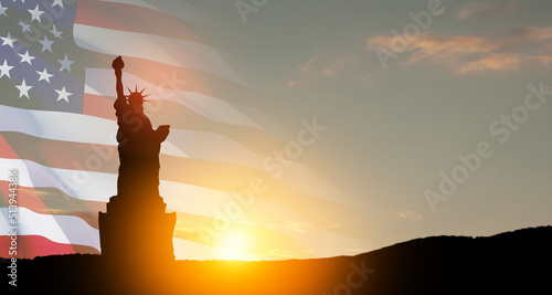 Statue of Liberty with a large american flag and sunset sky on background.