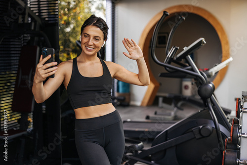 Pretty young caucasian woman communicates by video link via smartphone standing in gym. Brunette wears sporty black top and leggings. Technology concept