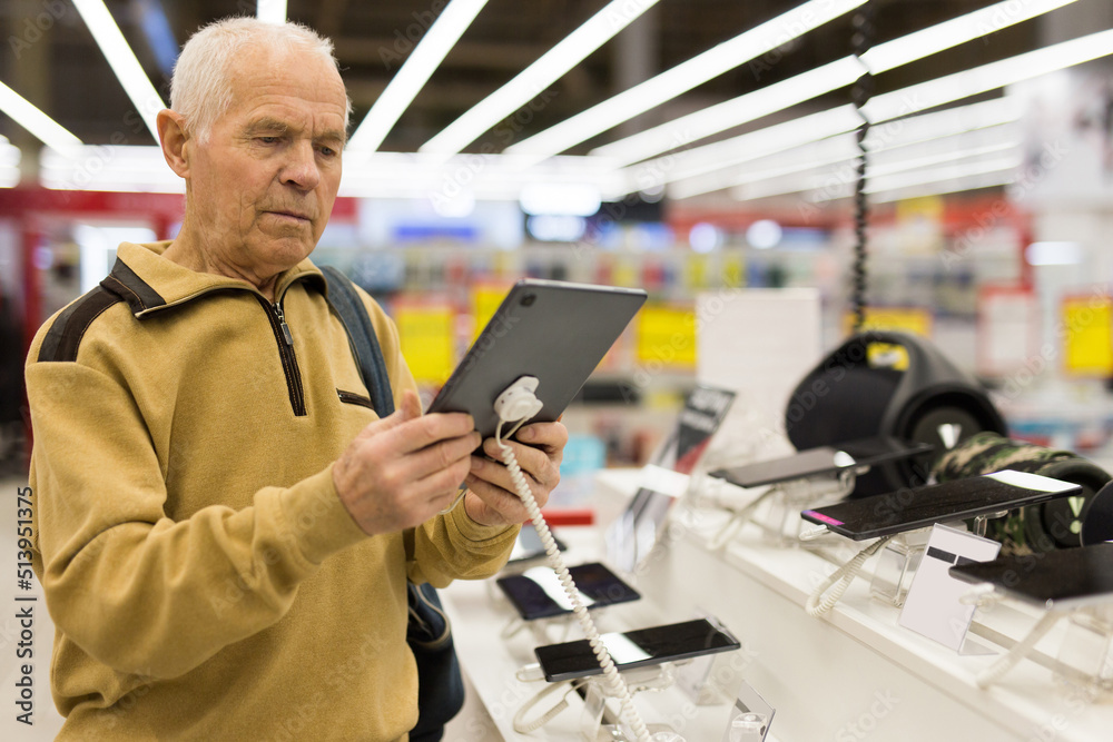 elderly man examines tablet computer in showroom of electronics store elderly man examines tablet computer in showroom of electronics store