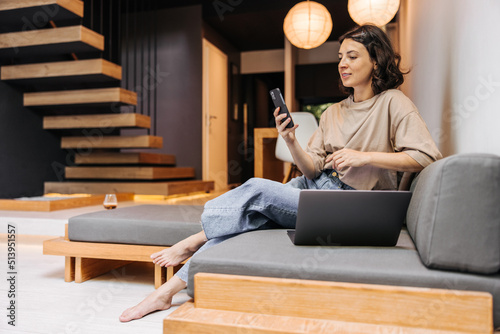Stylish young caucasian woman is typing message on smartphone sitting on sofa. Brunette is browsing site on mobile phone. Technology concept