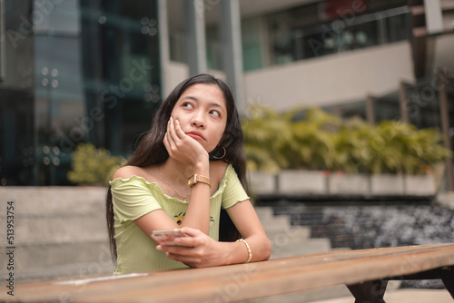 A bored or lonely young asian woman staring blankly sitting at the bench near the mall. Having no friends and feeling down. photo