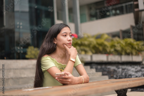 A bored or lonely young asian woman staring blankly sitting at the bench near the mall. Having no friends and feeling down.