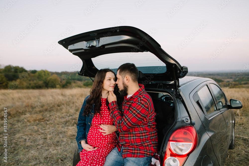 Couple of future young parents sit in the trunk of their family car and hug. Red palette, countryside outside the city, the autumn landscape, the concept of a happy pregnancy and future motherhood
