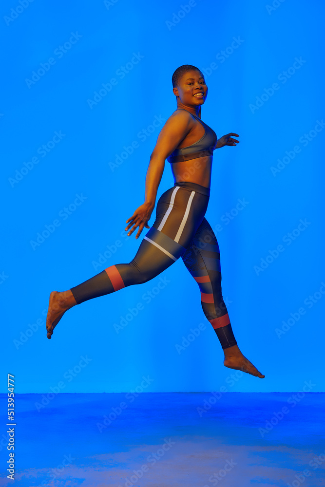 Happy african american woman wearing sportive clothes showing plus size hips. Body positive concept. Blue light on background Happy african american woman wearing sportive clothes showing plus size hips. Body positive concept. Blue light on background