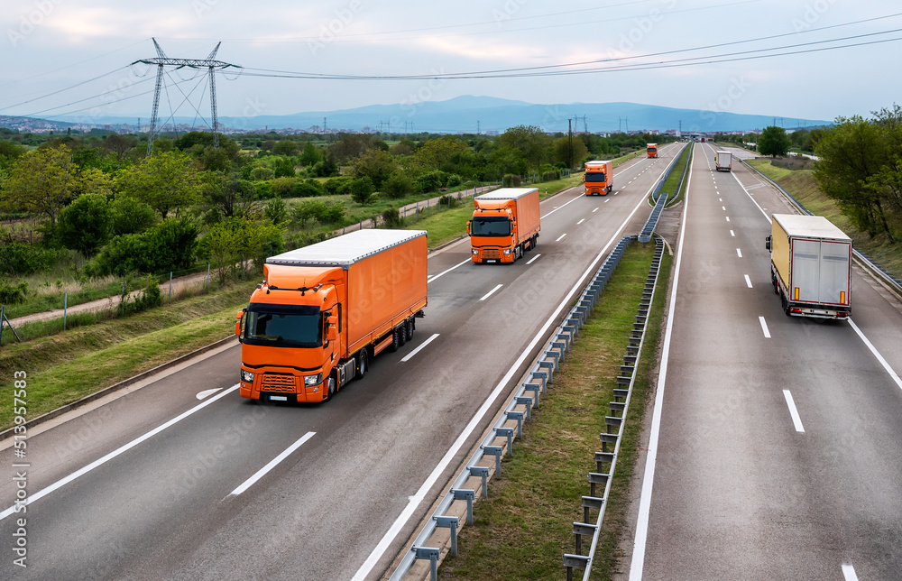 Four lanes Highway Transit with orange and yellow transportation trucks on a bright blue day. Highway traffic. Four lanes Highway Transit with orange and yellow transportation trucks on a bright blue day. Highway traffic.