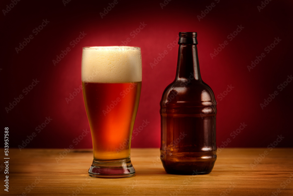 Glass of beer and a bottle on a wooden table and dark red background. Craft beer of local brewery. Focus is on the glass with beer.