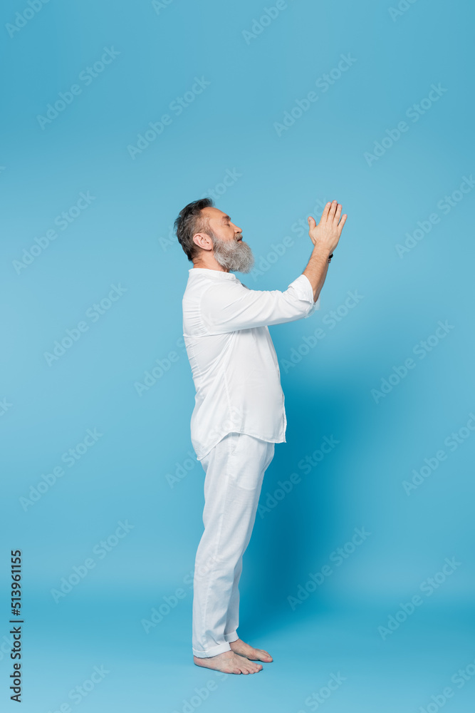 side view of senior yoga master meditating with praying hands on blue background.