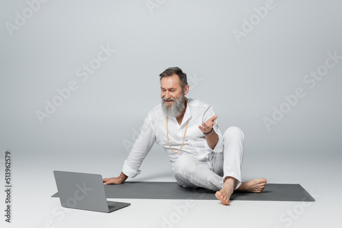 guru mentor sitting on yoga mat with crossed legs and talking near laptop on grey.