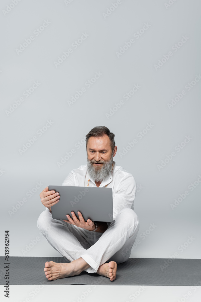 senior yoga coach with laptop sitting on yoga mat in easy pose on grey. senior yoga coach with laptop sitting on yoga mat in easy pose on grey.