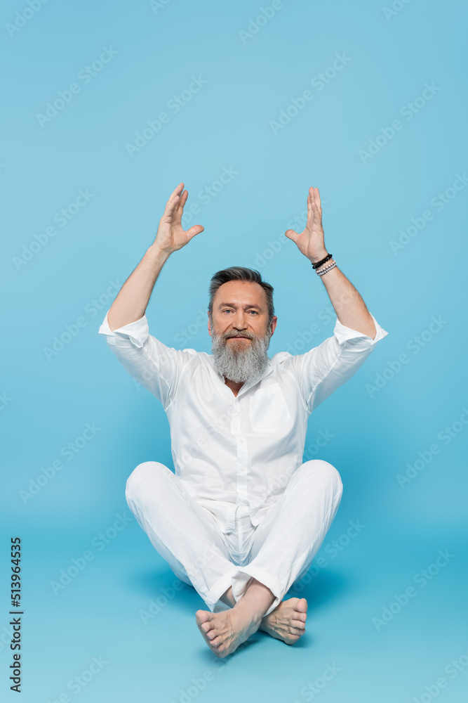senior yoga master looking at camera while meditating in easy pose with raised hands on blue. senior yoga master looking at camera while meditating in easy pose with raised hands on blue.