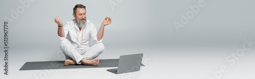 senior yoga coach showing chin mudra near laptop while sitting in easy pose on grey, banner.