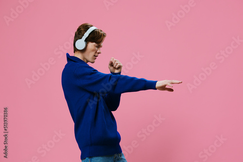 funny young student pretends to sing while listening to music in headphones while standing in a blue sweater on a pink background