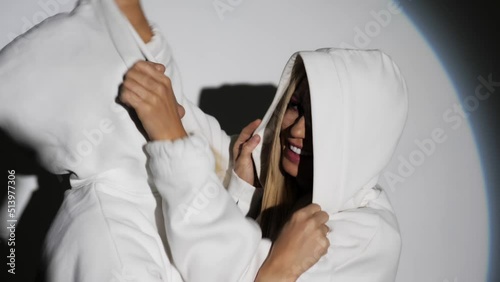 Two blondes with bright makeup in white hoodies have fun on camera. Studio with a white background, illuminated by the light from the spotlight.