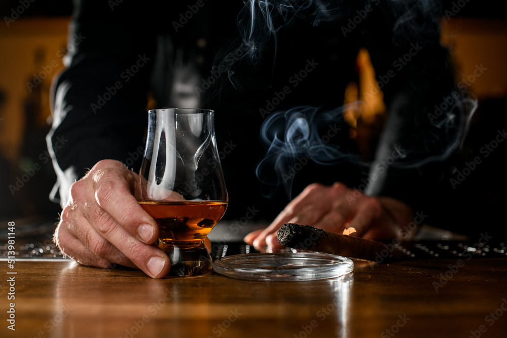 male hand holds glass with cognac and ashtray with a smoking cuban cigar nearby