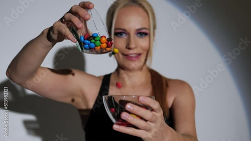 A blonde with bright makeup pours multicolored candies from a glass cup into another bowl.Slow motion, White background, studio lighting.