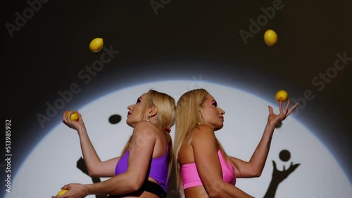 Blondes juggle lemons, standing with their backs to each other, slow motion.The girls are illuminated by a spotlight, with a white background behind.