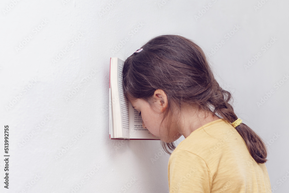 Sad and tired caucasian girl with dyslexia holds a book with her forehead. The child learns to speak and read correctly - obrazy, fototapety, plakaty 