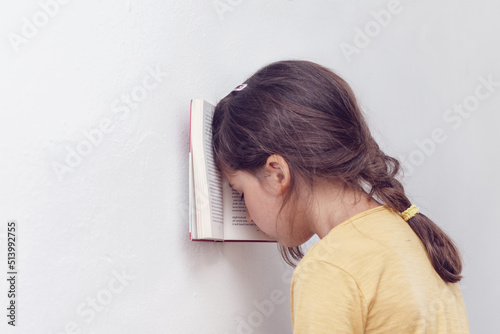 Sad and tired caucasian girl with dyslexia holds a book with her forehead. The child learns to speak and read correctly