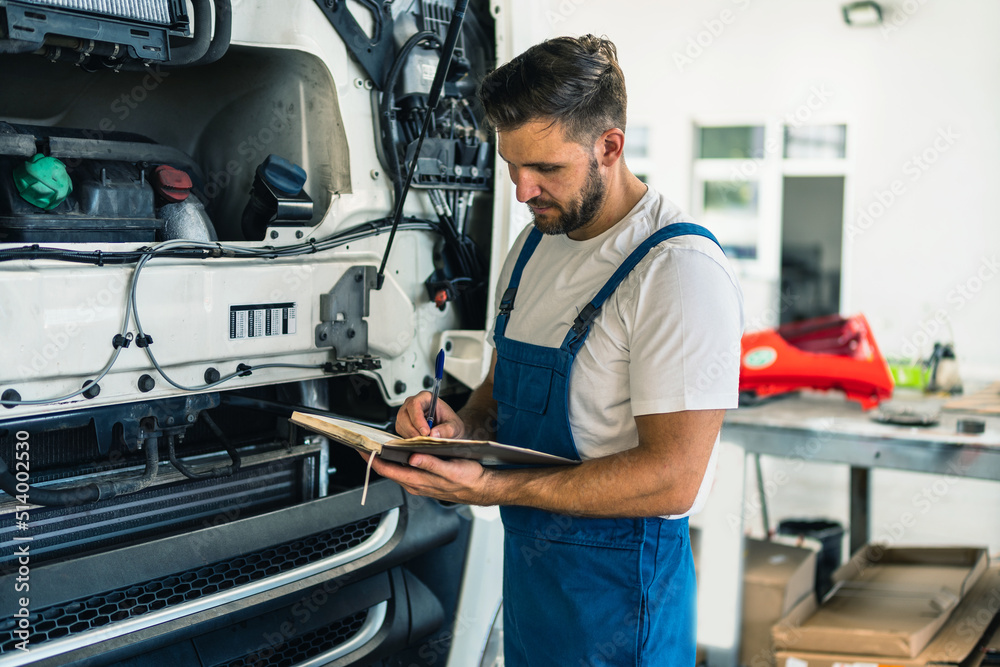 Truck mechanic writing notes in notebook during working in modern workshop. Young concentrated caucasian bearded man.Truck service, repair. Garage interior with tools and equipment Truck mechanic writing notes in notebook during working in modern workshop. Young concentrated caucasian bearded man.Truck service, repair. Garage interior with tools and equipment