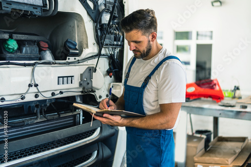 Truck mechanic writing notes in notebook during working in modern workshop. Young concentrated caucasian bearded man.Truck service, repair. Garage interior with tools and equipment