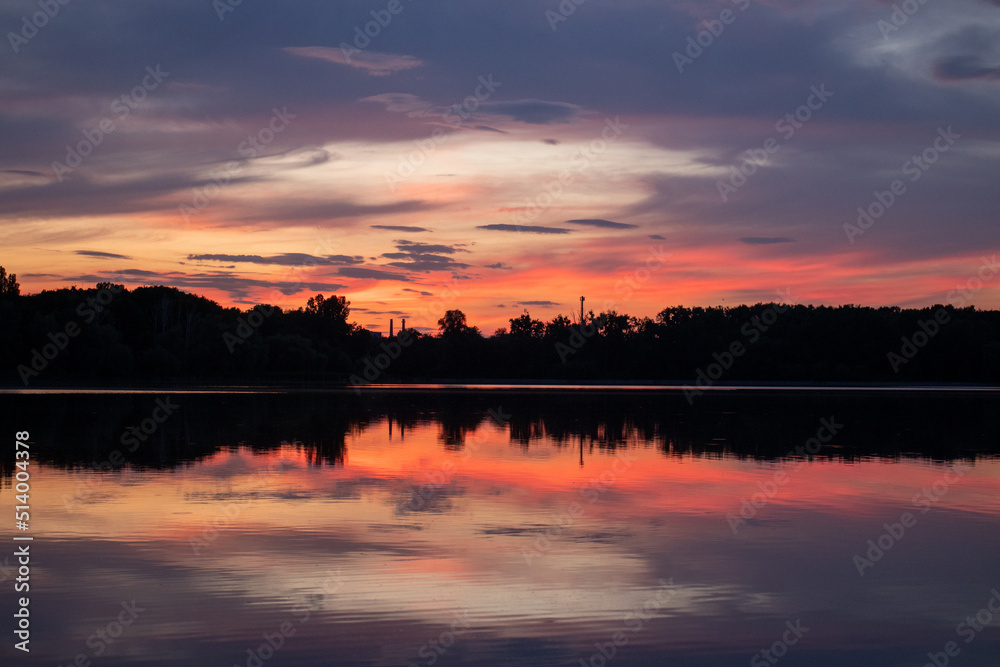 Sunset over the river. Scenic summer landscape with trees silhouettes and clouds reflections in the water