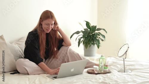 Young redhead woman uses laptop and laughs while lying on bed. Cosmetics at home, online learning, remote work, caucasian female freelancer in pandemic covid 19. 4k footage