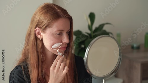 Close-up of redhead woman using jade roller on face looking in the mirror. Rejuvenation treatment. Red hair young girl doing beauty routine at home. Concept of skincare, cosmetics, wellbeing. 4k