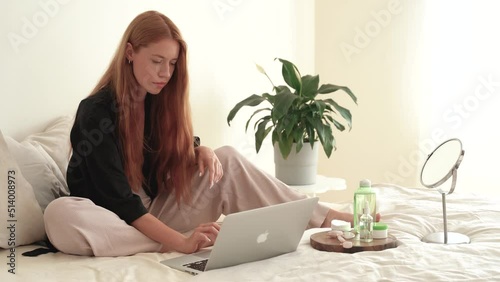 Young redhead woman uses laptop and drinking from large cup on bed. Cosmetics at home, online learning, remote work, caucasian female freelancer in pandemic covid 19. 4k footage