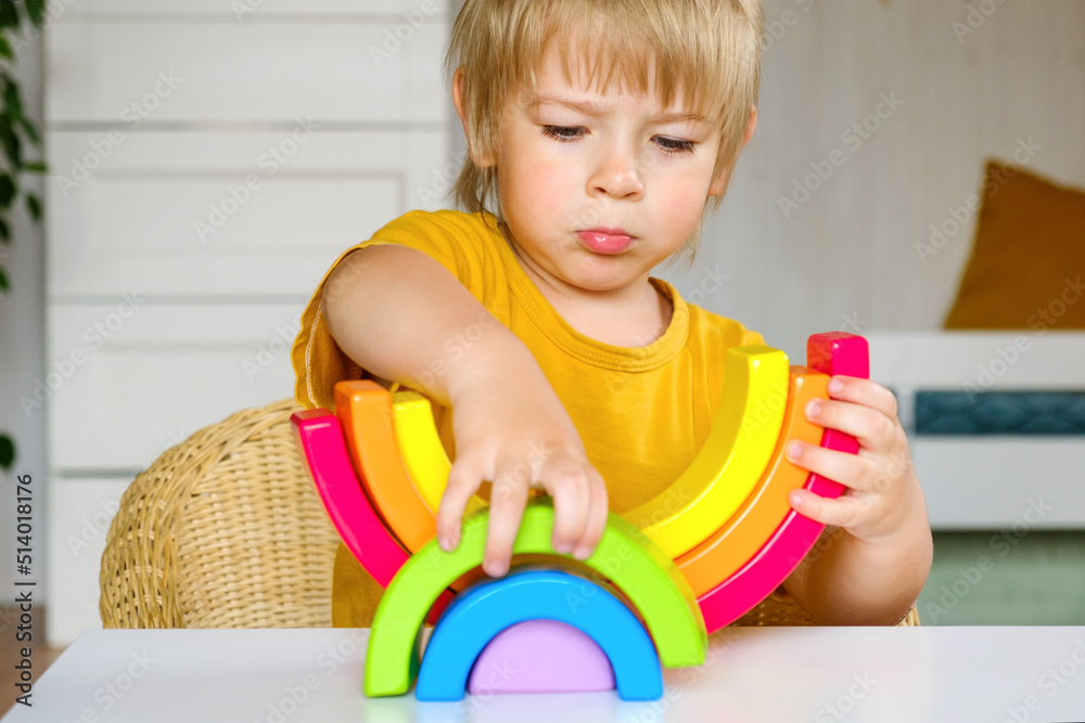 Little blond hair boy in clothes made of natural fabric plays with rainbow colored wooden toys at white table. Cozy room background in scandinavian style. Warm natural colors. Eco friendly lifestyle Little blond hair boy in clothes made of natural fabric plays with rainbow colored wooden toys at white table. Cozy room background in scandinavian style. Warm natural colors. Eco friendly lifestyle