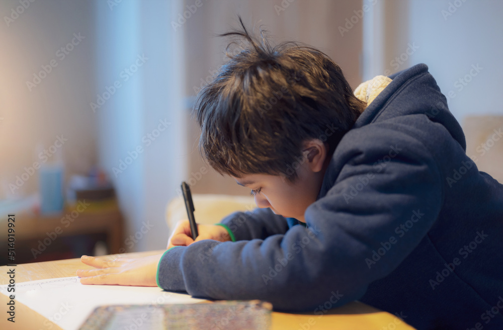 Kid siting on table doing homework,Child boy holding black pen writing on white paper,Young boy practicing English words at home. Elementary school and home schooling, Distance Education concept Kid siting on table doing homework,Child boy holding black pen writing on white paper,Young boy practicing English words at home. Elementary school and home schooling, Distance Education concept