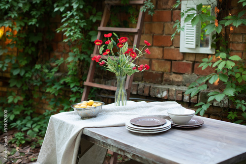 A bouquet of red poppies in a glass vase. Flowers in a vase at home. Interior Terrace, patio. Wooden table and plates for garden party or dinner. Table for lunch outside in garden in yard of house.	 photo