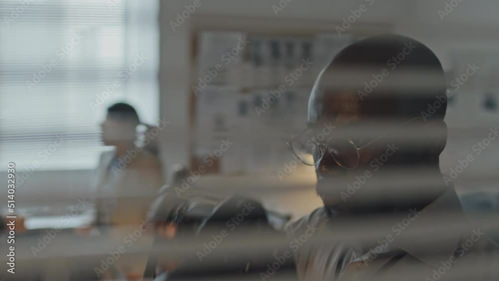 Chest-up view through window with horizontal blinds of young African American male agent taking pictures on professional camera from cover in office Chest-up view through window with horizontal blinds of young African American male agent taking pictures on professional camera from cover in office