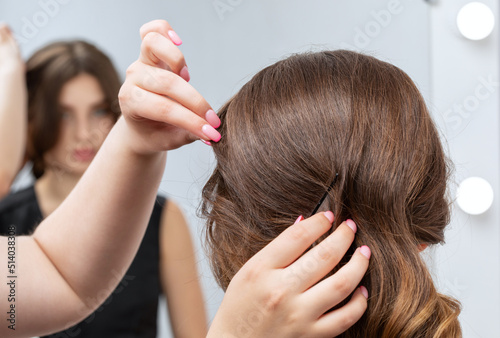 The hairdresser makes a hairstyle to a young attractive woman with a beautiful make-up in a black dress in a beauty salon.