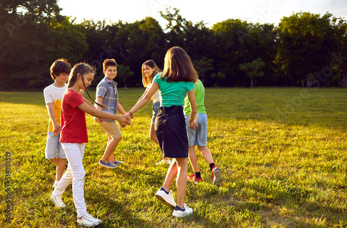 Fototapeta Naklejka Na Ścianę i Meble -  Cheerful playful preteen children in sunny summer park walking in circle holding hands. Boys and girls in casual clothes having fun outdoors making circle to play together. Childhood concept.