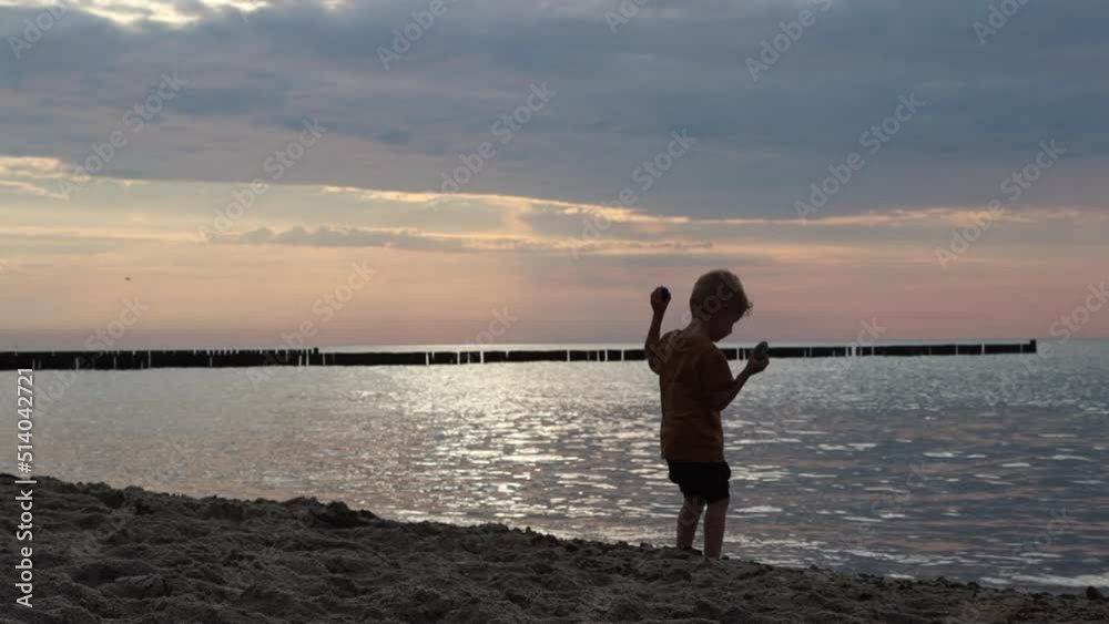 A little boy confidently walks along the sand to the sea and throws stones into the water in the evening at sunset A little boy confidently walks along the sand to the sea and throws stones into the water in the evening at sunset