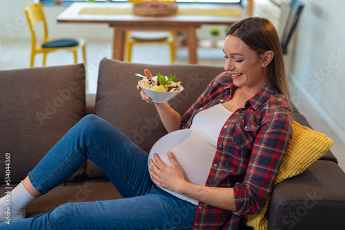 Beautiful happy pregnant young woman sitting and eating fruit salad on sofa at home