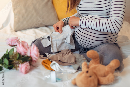 Pregnant woman holding baby bodysuit and packing maternity hospital bag. Beautiful mother during pregnancy waiting for baby preparing suitcase of clothes  toy and necessities for newborn child birth.