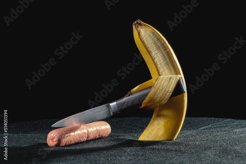 A ripe banana cuts a sausage with a knife on a stone tabletop with forks on a black background.