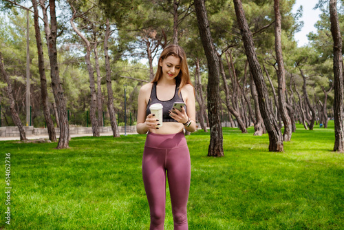 Cute caucasian woman wearing sportive clothes on city park, outdoors holding takeaway coffee mug and using mobile smart phone. Outdoor sports, healthy life concepts. © platinumArt