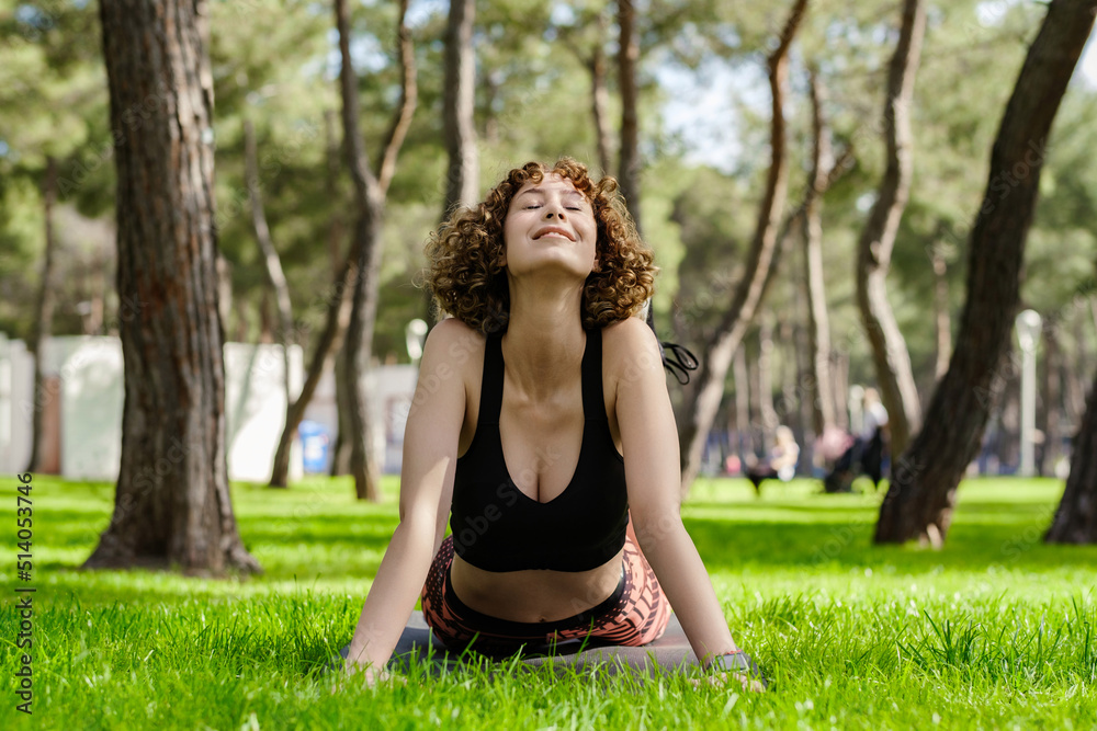 Young caucasian woman wearing black sports bra standing on city park, outdoor doing yoga, stretching on yoga mat. Practicing stretching high cobra asana pose.