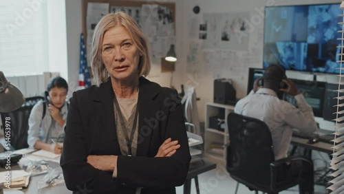 Waist-up portrait of mature Caucasian female prosecutor in formal suit crossing arms on chest, standing in office at daytime, looking on camera
