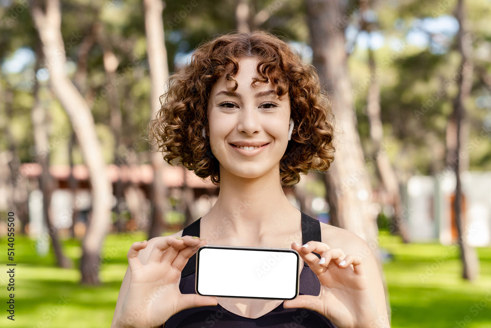 Young redhead girl smiling happy wearing sports bra standing on city park, outdoors showing blank smartphone screen. Fitness app. Recommending new mobile application, mockup. Young redhead girl smiling happy wearing sports bra standing on city park, outdoors showing blank smartphone screen. Fitness app. Recommending new mobile application, mockup.