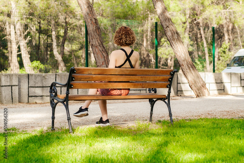 Beautiful redhead woman wearing black sports bra standing on city park, outdoors sitting on a bench and watching. Spending time alone. Thinking about life. Peaceful atmosphere. Back view.