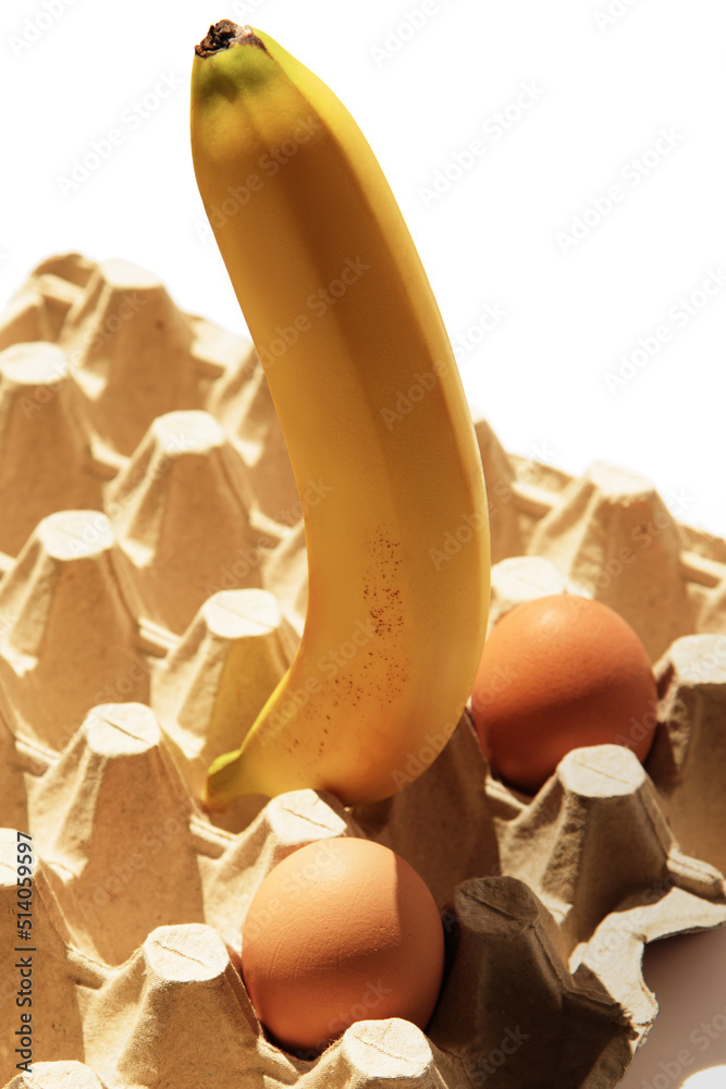 Big ripe banana and two chicken eggs in egg carton on white background in natural sunlight. Sexy concept.