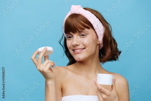 a joyful, happy woman, wrapped in a towel, stands on a blue background holding a white jar of cream in her hand, holding the lid in her hand and smiling, squinting her eyes